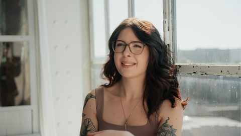 Junge Frau mit ENNY Brille im Cat-Eye-Stil sitzt entspannt am Fenster, lächelt in die Sonne. Stilvolle Brillenmode mit Persönlichkeit.
