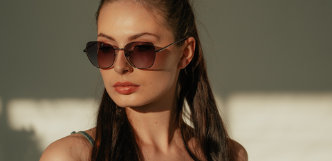 Frau mit langen dunklen Haaren trägt elegante Sonnenbrille mit dunkel getönten Gläsern und filigranem Metallrahmen, schaut seitlich in warmes Licht mit weichen Schatten im Hintergrund.