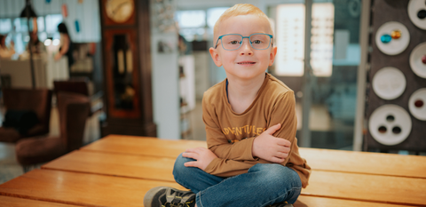 Kleiner Junge mit blonden Haaren und hellblauer Kinderbrille sitzt lächelnd auf einem Holztisch in einem modernen Optikgeschäft, trägt braunes Shirt mit „Adventure“-Aufdruck.