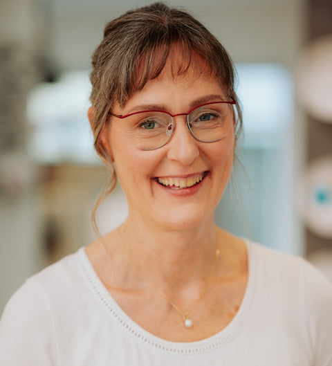 Lächelnde Frau mit grauem Haar und weißem Oberteil trägt die ENNY Damenbrille Heda in Rot-Silber mit eleganter Schmetterlingsform; das Tragefoto zeigt die Brille im Alltagseinsatz mit natürlichem Ausdruck im Optikerambiente