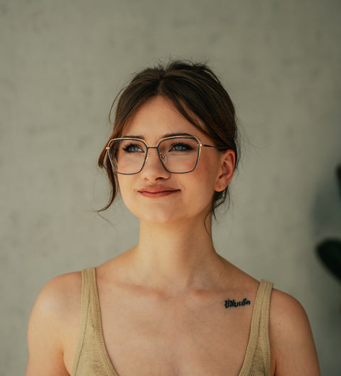 Frau mit lockerem Dutt und Tattoo auf der Schulter trägt die eckige ENNY Juna Damenbrille aus Titan in Dunkelblau mit glänzenden Roségold-Details vor hellem Hintergrund.