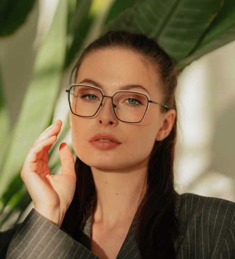 Frau mit Nadelstreifenblazer und offenem Haar trägt die eckige ENNY Juna Damenbrille aus Titan in mattem Schwarz mit weißen Details vor großem Blattgrün