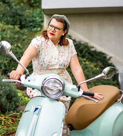 Frau mit Brille auf einer türkisen Vespa, gekleidet in ein Blümchenkleid – perfekter Retro-Style.