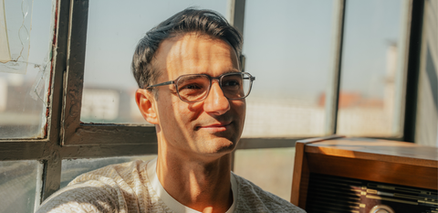 Mann mit moderner, transparenter Brille sitzt entspannt am Fenster im warmen Sonnenlicht.