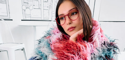 Junge Frau mit auffälliger bunter Kunstfelljacke und dazu passender Kunststoffbrille in rot sitzt vor einem stilisierten schwarz-weiß Hintergrund im Comic-Stil