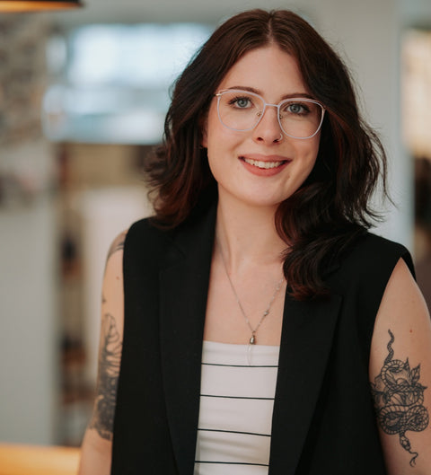 Junge Frau mit dunklen Haaren trägt die ENNY Damenbrille Sarah in zartem Flieder mit roséfarbenen Details – moderne Schmetterlingsform, stilvoll kombiniert mit schwarzer Weste und weißem Top, aufgenommen im Optikerfachgeschäft
