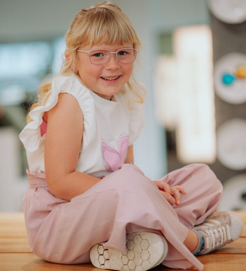 Fröhliches Mädchen mit blonden Haaren trägt die runde Kinderbrille ENNY Waldi in Pink-Rosa – verspielter Look mit zarter Brillenfassung, aufgenommen beim Optiker auf einer Holzbank sitzend in entspannter Pose