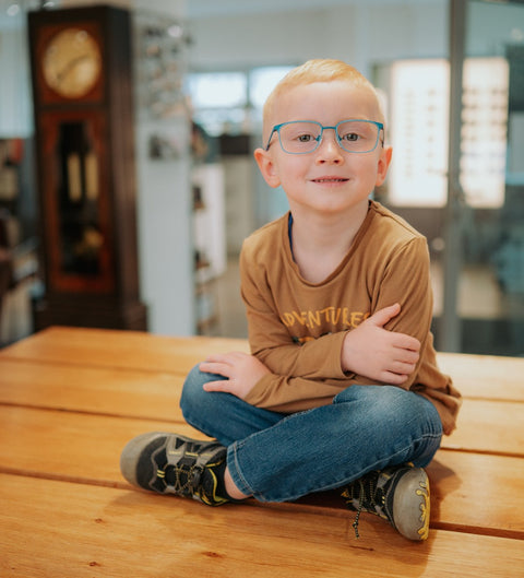 Junge mit blonden Haaren und blauer, eckiger Kinderbrille ENNY Waldi sitzt lächelnd im Schneidersitz auf einer Holzbank beim Optiker – fröhliches Tragebild in heller Umgebung