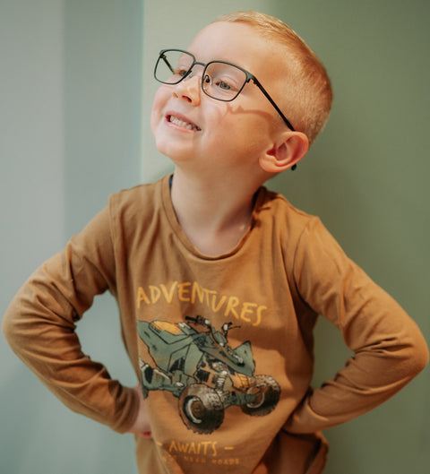 Lachender Junge mit dunkelgrüner, eckiger Kinderbrille ENNY Waldi posiert selbstbewusst mit Händen in der Hüfte vor mintgrünem Hintergrund – modernes Tragebild beim Optiker