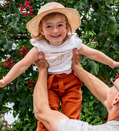 Fröhliches kleines Mädchen mit rosa ENNY Kinderbrille Lina und Strohhut wird spielerisch von einem Erwachsenen in die Luft gehoben, im Hintergrund blühender Baum