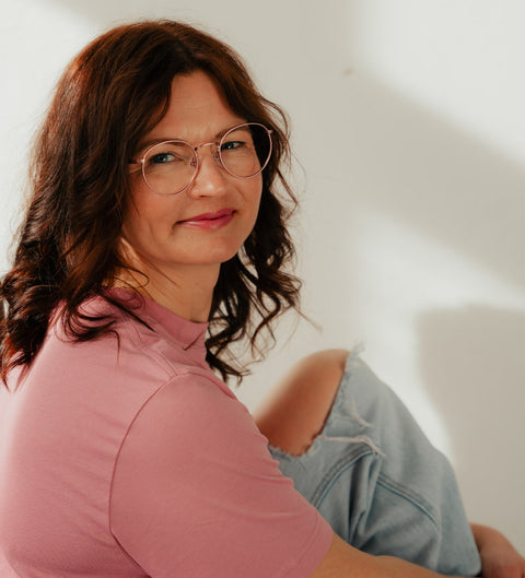 Frau mit rötlich-braunen Locken trägt die ENNY Brille „Sico“ in metallischem Rosa, rundes Modell im soften Sonnenlicht, gemütlicher Look mit T-Shirt