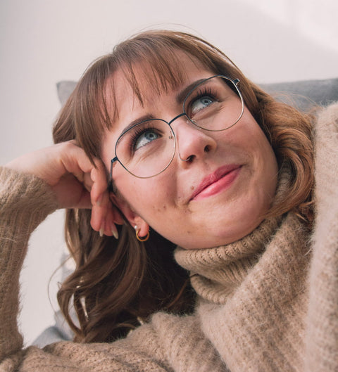 Frau mit runder ENNY Damenbrille in Farbverlauf Petrol-Gold, gemütlich liegend auf einem Sofa in einem beigen Rollkragenpullover
